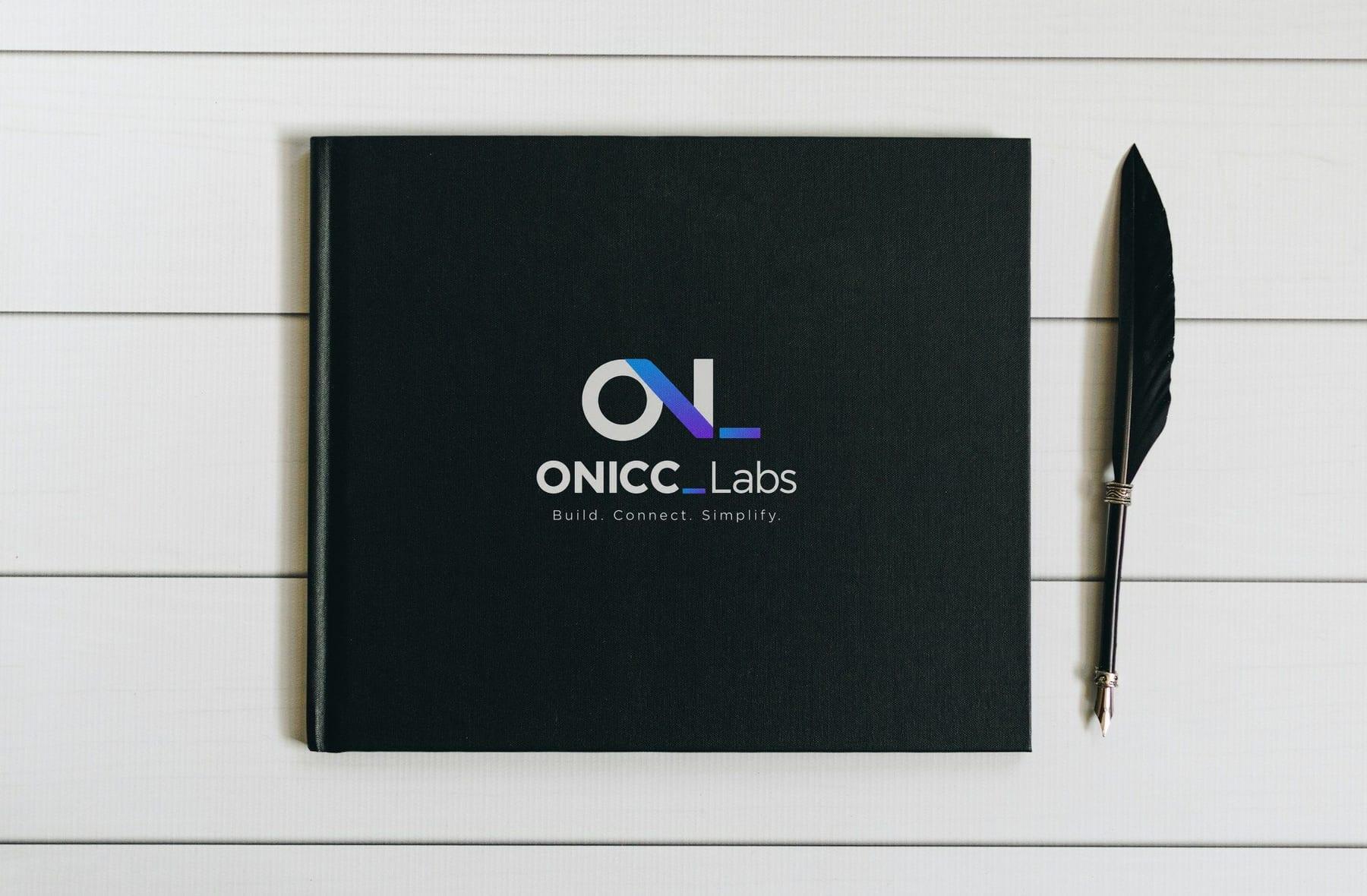 Black hardcover diary with the ONICC Labs logo printed on the cover, beside a black quill pen