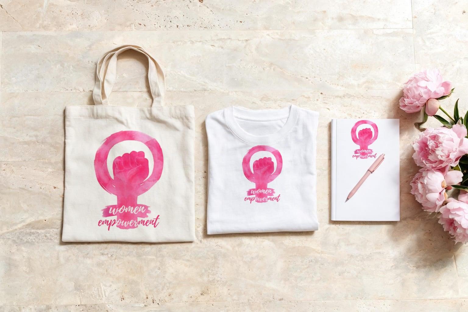 Off-white canvas tote, white T-shirt and white notebook, each printed with a pink watercolour women empowerment mark, on pale stone beside pink peonies