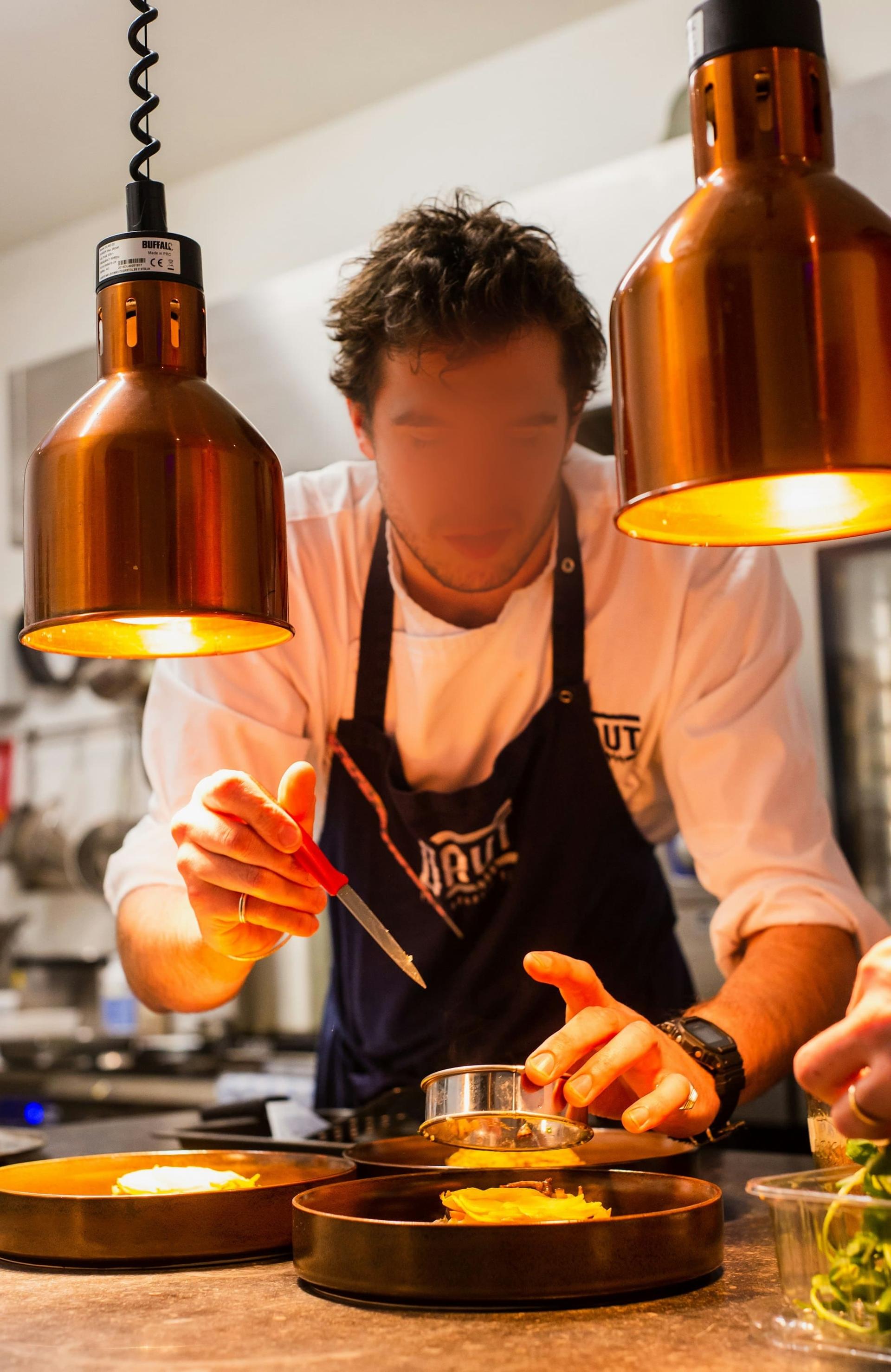A chef finishing plates under copper heat lamps
