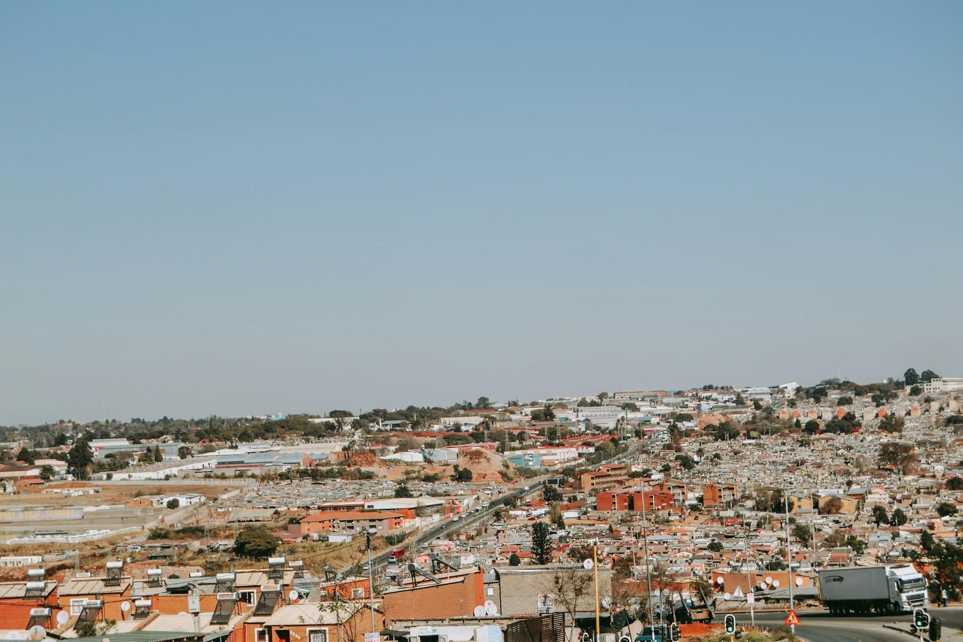 Aerial view across low, closely packed rooftops stretching to the horizon under a hazy Highveld sky