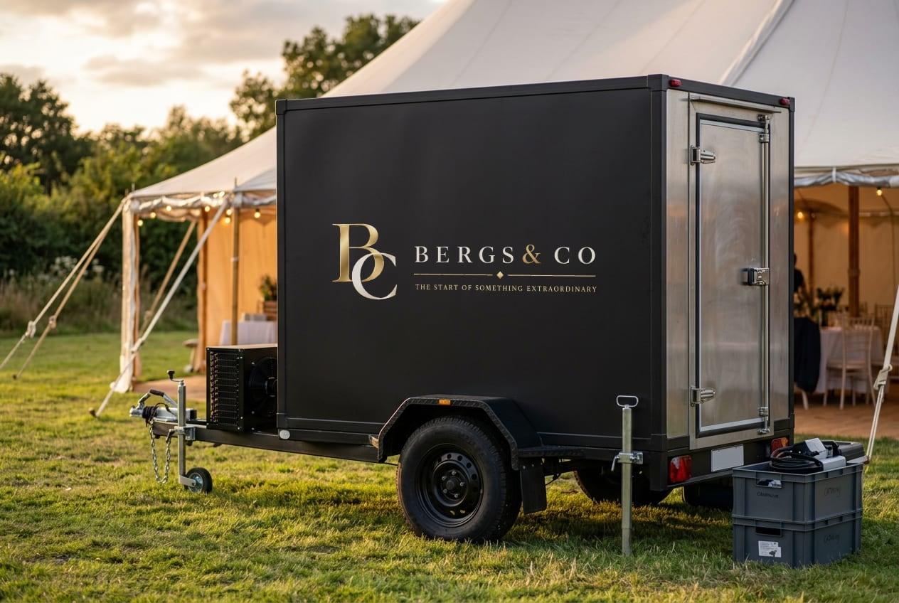 Black single-axle mobile freezer trailer beside a marquee at golden hour, its side panel carrying the gold Bergs & Co lockup