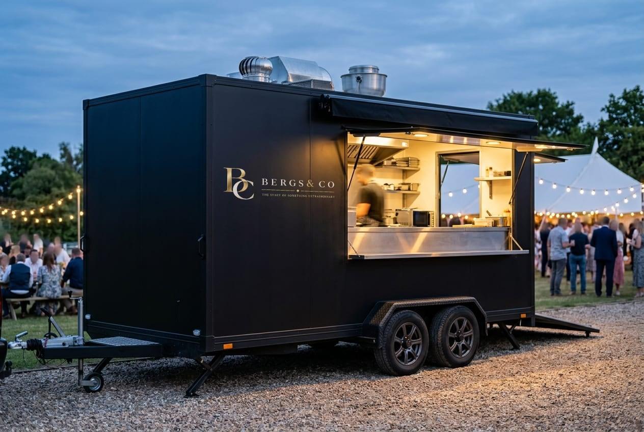 Black mobile kitchen trailer at dusk with its lit serving hatch open and the gold Bergs & Co lockup on its side panel